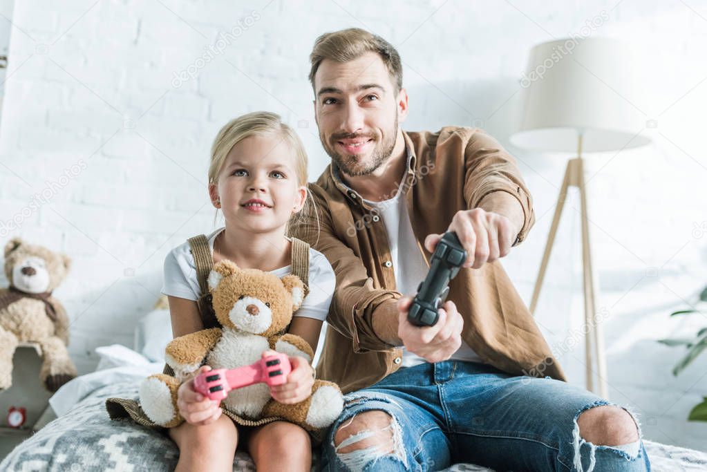 happy father and cute little daughter with teddy bear playing with joysticks at home