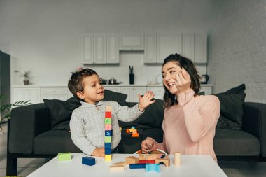 küçük bir çocuk ve blok ahşap kule oyun evde oynarken annesi gülümseyerek