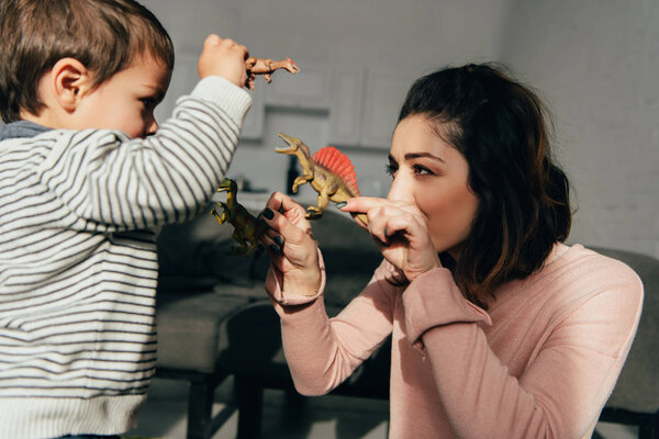selective focus of mother and little son playing toy dinosaurs in living room at home