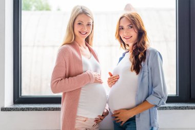 pencere duran ve kameraya bakarak gülümseyen hamile kadınlar 