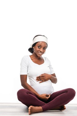 happy african american pregnant woman sitting on floor and keeping hands on belly isolated on white clipart