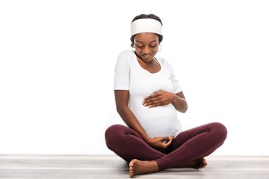 african american pregnant woman sitting on fitness mat and looking on belly isolated on white clipart