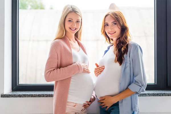 Leende Gravida Kvinnor Står Nära Fönstret Och Tittar Kameran — Stockfoto