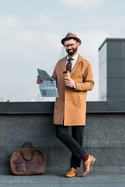 Uśmiechający Się Brodaty Biznesmen Stojąc Skrzyżowanymi Nogami Trzymając Gazetę Kawy — Darmowe zdjęcie stockowe