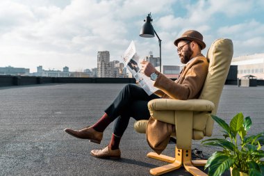 bearded businessman sitting in armchair and reading newspaper on roof  clipart