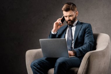 dizüstü bilgisayar ile koltuk oturan ve smartphone üzerinde konuşurken kendinden emin işadamı 