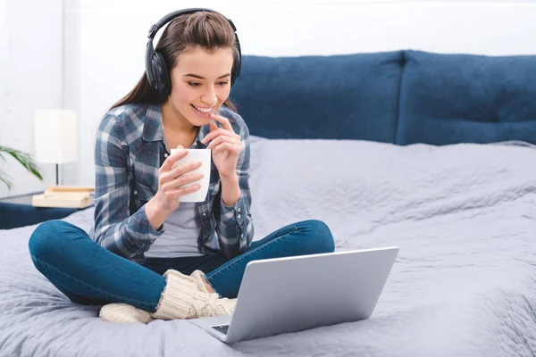 Glad Ung Kvinna Hörlurar Håller Cup Och Använder Laptop Säng — Stockfoto