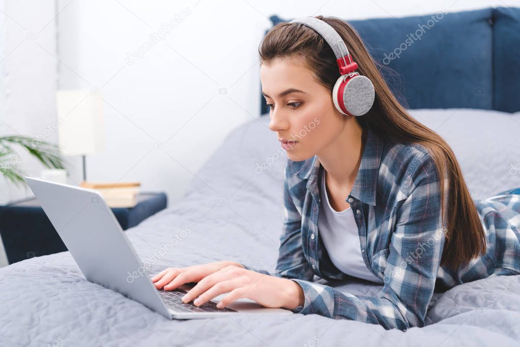 beautiful young woman in headphones using laptop and lying on bed
