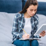 Young woman holding digital tablet and writing in notebook in bedroom