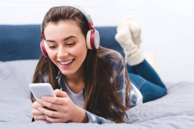 güzel gülümseyen genç kadın yatakta yatarken ve smartphone kullanarak kulaklık