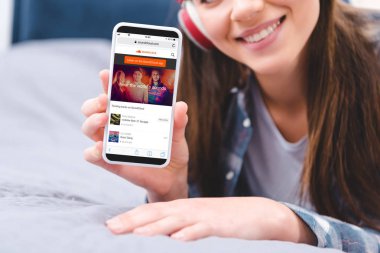 gülümseyen genç kadın yatakta yatarken soundcloud uygulama ile akıllı telefon tutarak Kulaklığımda kırpılmış atış 