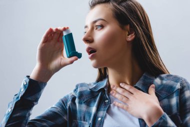 young woman using asthma inhaler and looking away isolated on grey clipart