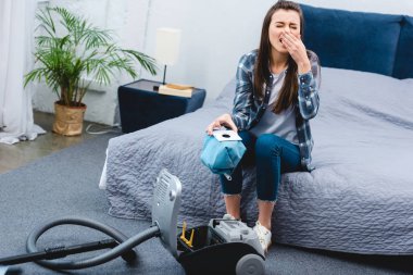 high angle view of woman with allergy holding container from vacuum cleaner with dust and sneezing in bedroom    clipart