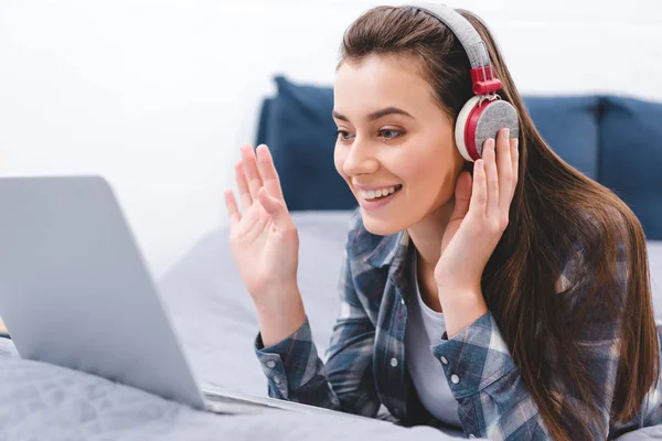 happy young woman in headphones using laptop and waving hand