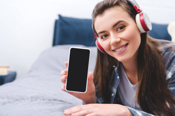 attractive girl in headphones holding smartphone with blank screen and smiling at camera while lying on bed 