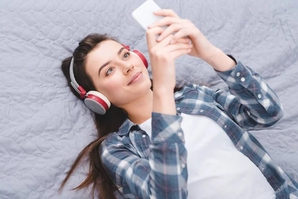 beautiful smiling girl in headphones lying on bed and using smartphone
