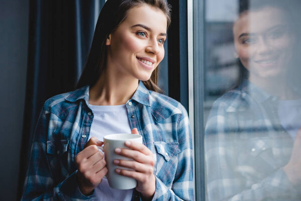 beautiful smiling young woman holding cup and looking at window  