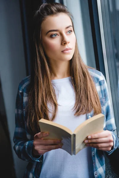 沉思的女孩在格子衬衫拿着书 看着窗口 — 图库照片