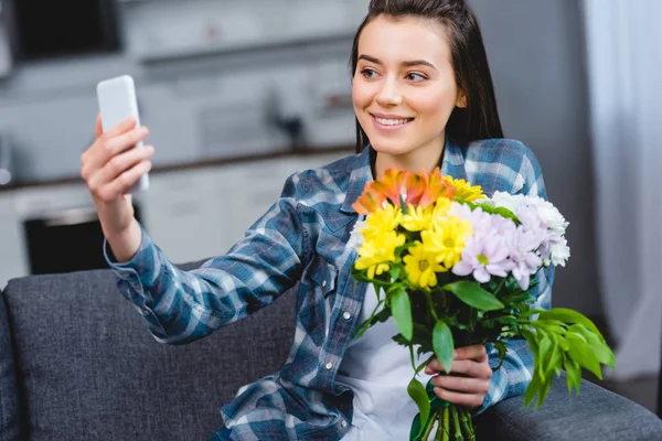 快乐的年轻女子拿着美丽的花朵 并采取自拍与智能手机在家里 — 免费的图库照片
