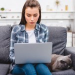 Menina usando laptop enquanto sentado no sofá com gato cinza bonito