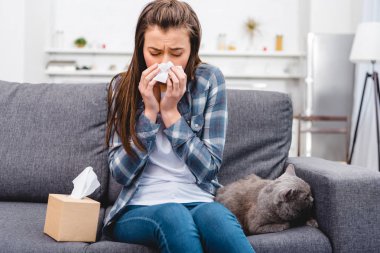 girl blowing nose in facial tissue while sitting with cat on couch clipart