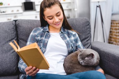 Genç kadın holding kitap gülümseyen ve kanepeye şirin gri kedi arıyorum