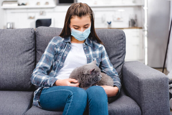 young woman in medical mask holding cat and suffering from allergy at home  