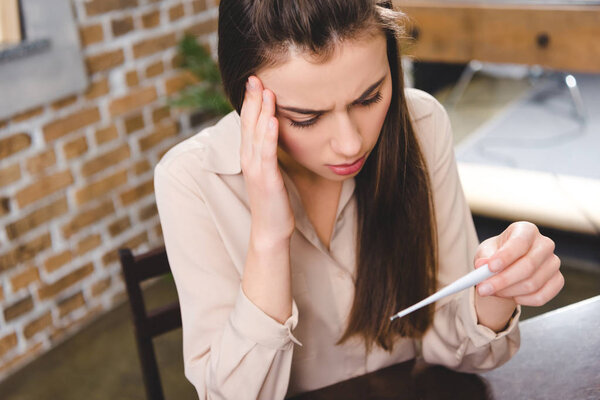 high angle view of sick young businesswoman holding thermometer and checking temperature in office 