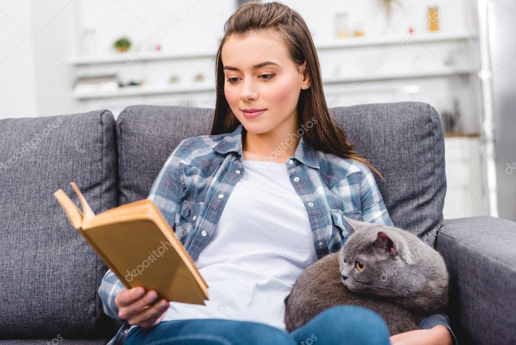 smiling young woman reading book and sitting with cat on couch