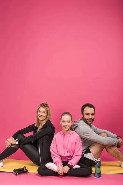 sportive family sitting on fitness mat with dumbbells and sports bottle, isolated on pink