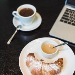 Plate with croissant and sauce, coffee and laptop on table in cafe