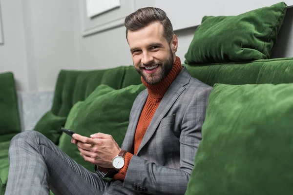 smiling handsome man in formal wear sitting on green sofa and using smartphone