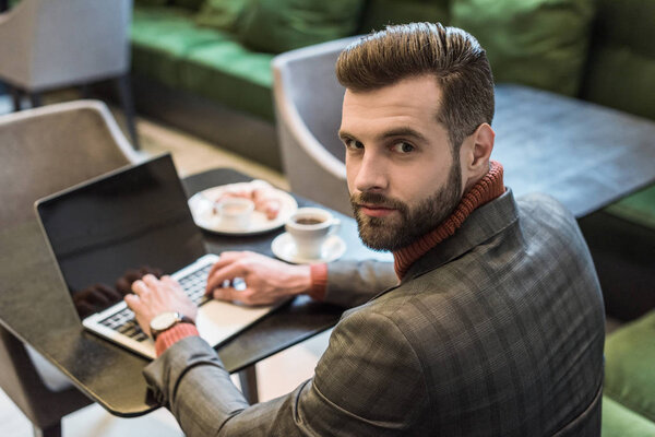 handsome businessman sitting at table, looking at camera and typing on laptop with blank screen in restaurant