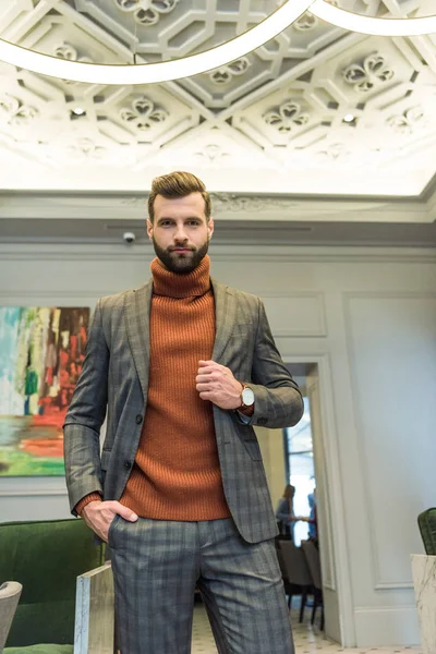 focused handsome man in formal wear looking at camera and posing with hand in pocket in lobby