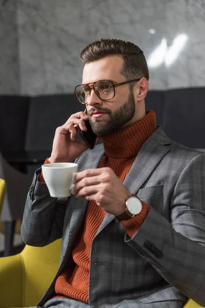 handsome man in formal wear and glasses talking on smartphone while drinking coffee in restaurant