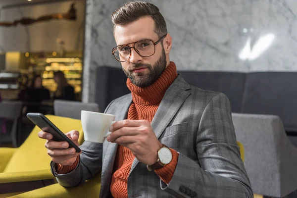handsome man in formal wear looking at camera, using smartphone and drinking coffee in restaurant