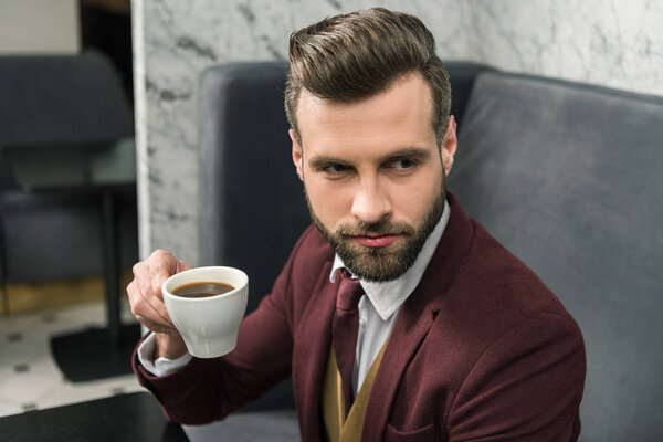 bearded handsome businessman in formal wear sitting at table and drinking coffee in restaurant