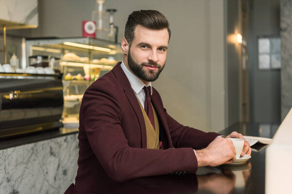 handsome businessman in formal wear looking at camera and sitting at table with coffee in restaurant