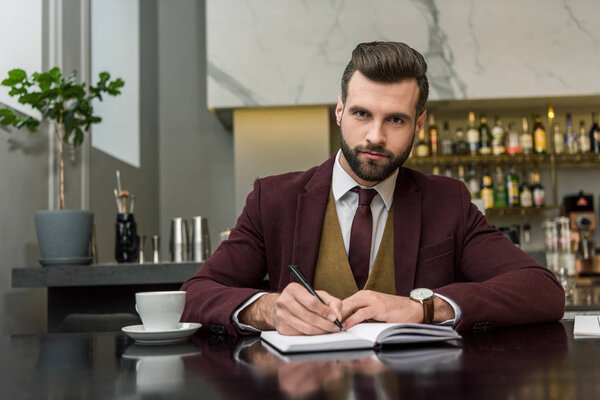 serious businessman in formal wear sitting and writing in notebook at table in restaurant