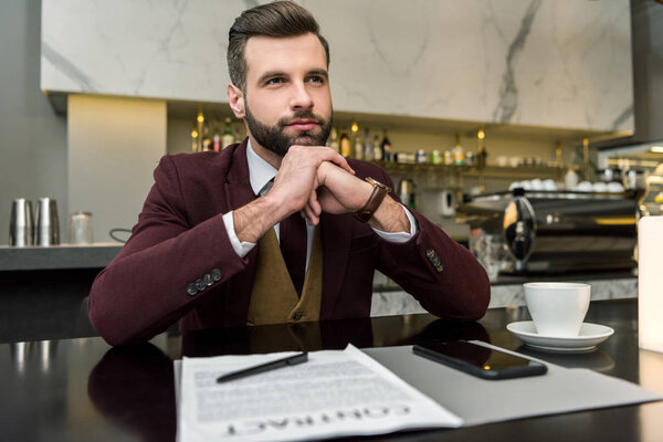 pensive businessman sitting at table with contract, coffee and smartphone while looking away