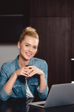 Kız holding bardak meyve suyu, kameraya bakarak ve laptop ile masada oturan gülümseyen