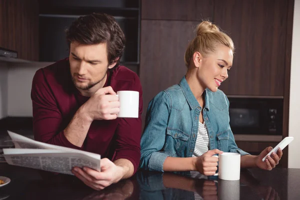 英俊的男人拿着咖啡杯和阅读报纸 而妻子使用智能手机在厨房 — 图库照片