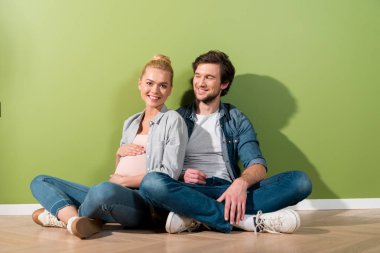 handsome man sitting on floor and looking at pregnant girl touching belly with both hands clipart