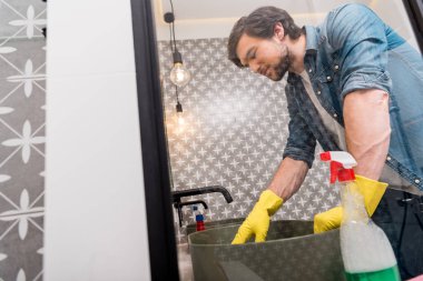 mirror reflection of handsome man in rubber gloves cleaning sink in bathroom clipart
