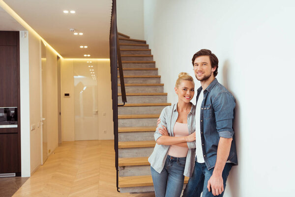 cheerful couple standing near stairs and looking at camera at home