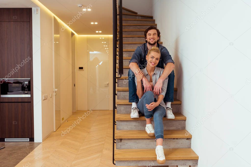 handsome man smiling, sitting on stairs and holding hands on shoulders of beautiful wife