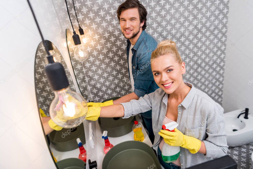 handsome man and attractive girl in rubber gloves girl cleaning mirrors and looking at camera in bathroom