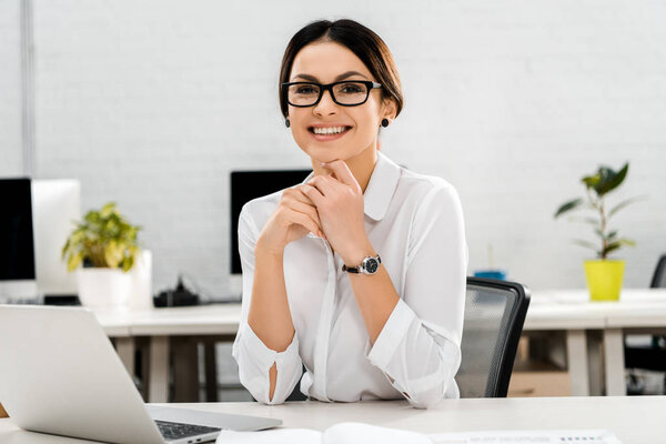 portrait of smiling businesswoman in eyeglasses at workplace with laptop in office