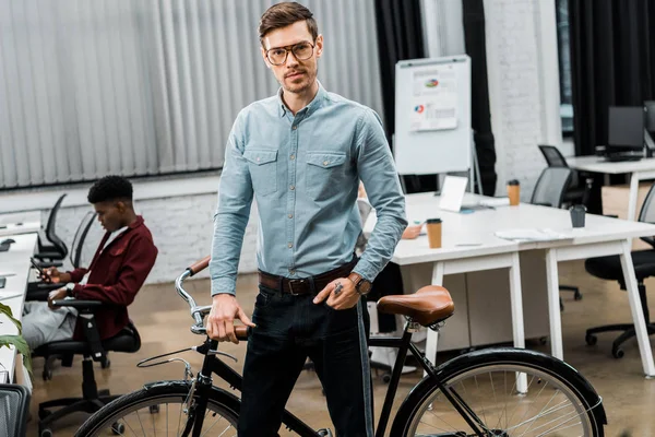 青年実業家の背後にあるアフリカ系アメリカ人の同僚とオフィスで自転車に傾いた  — 無料ストックフォト