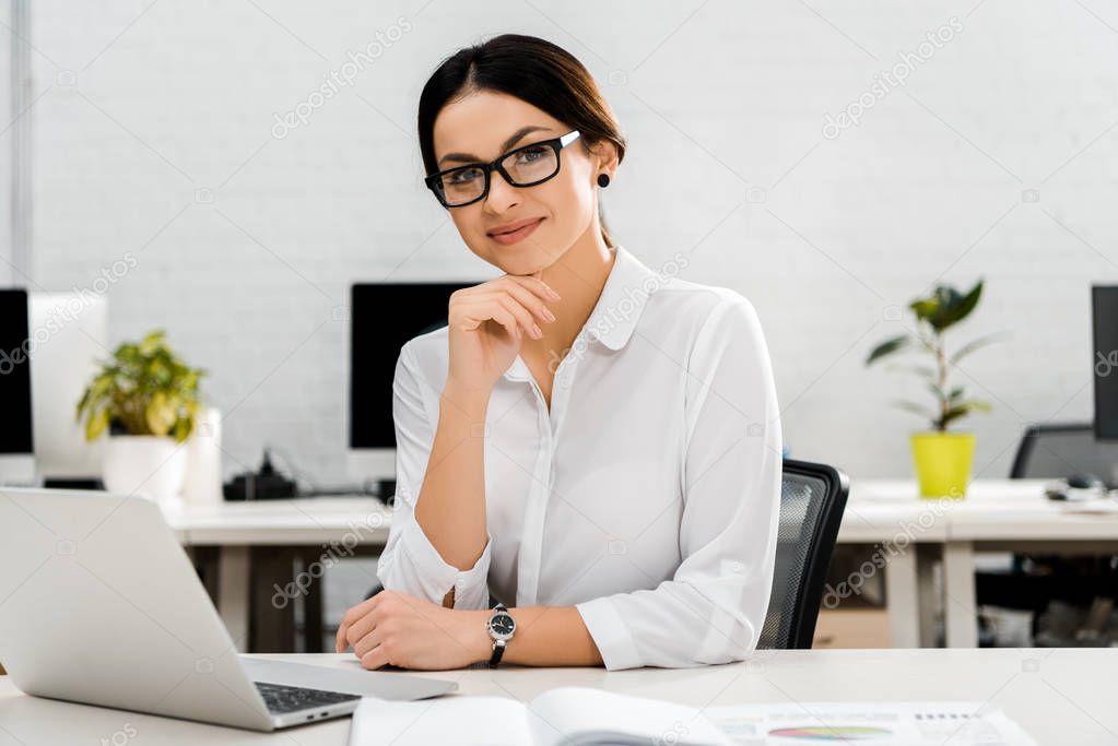 portrait of smiling businesswoman in eyeglasses at workplace with laptop in office